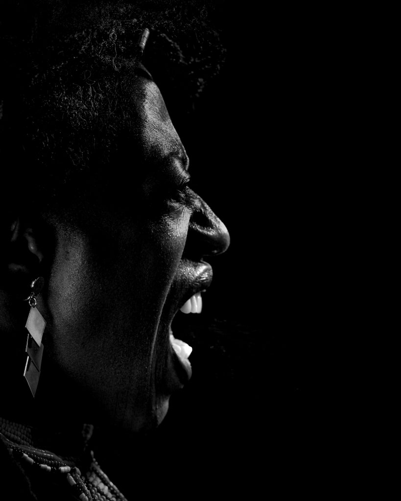 A black and white close-up of a person shouting, side profile. Dramatic lighting highlights intense emotion. Large earrings and textured hair are visible.