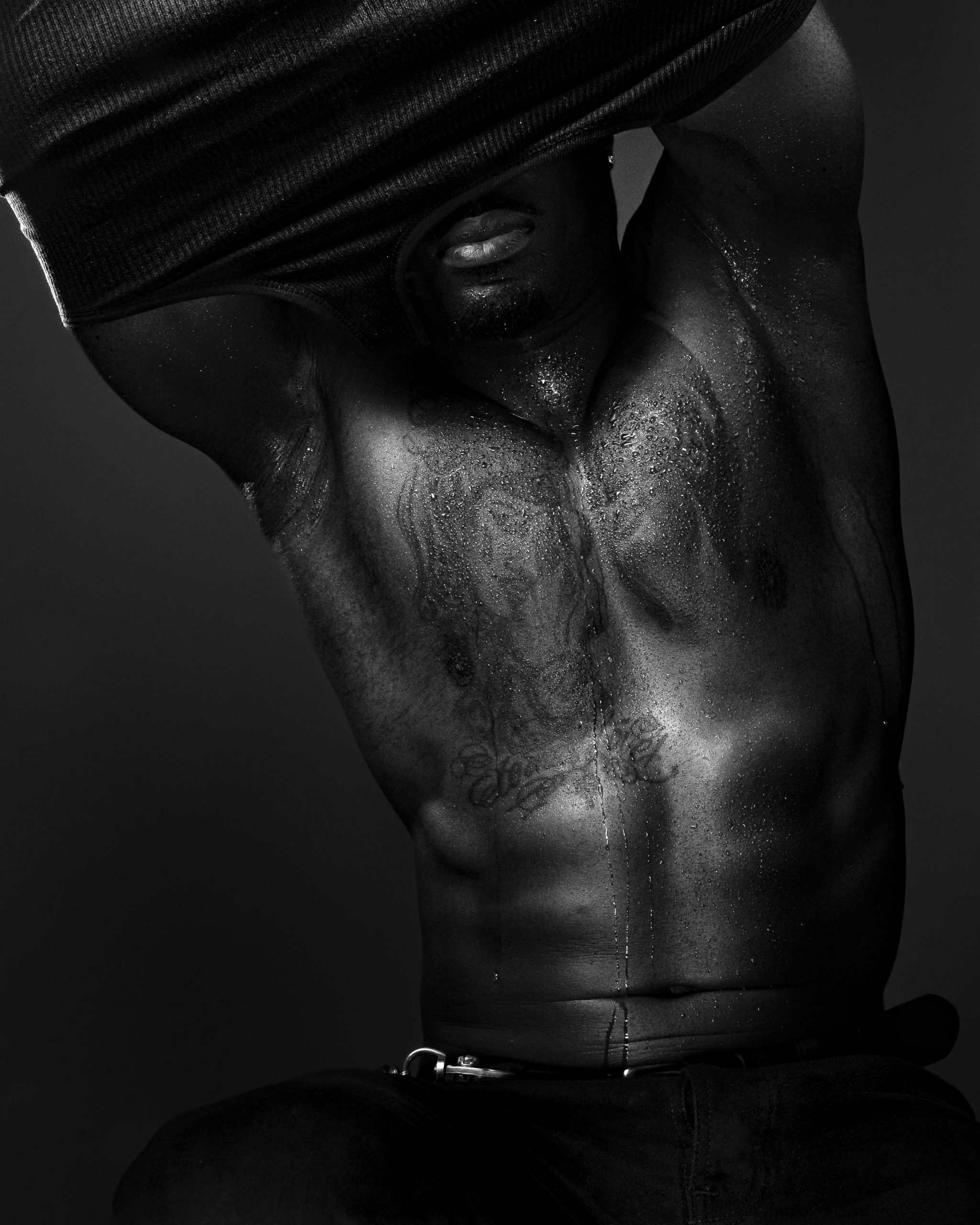 Black and white side profile of a tattooed man lifting his vest over his head to show his muscular torso.