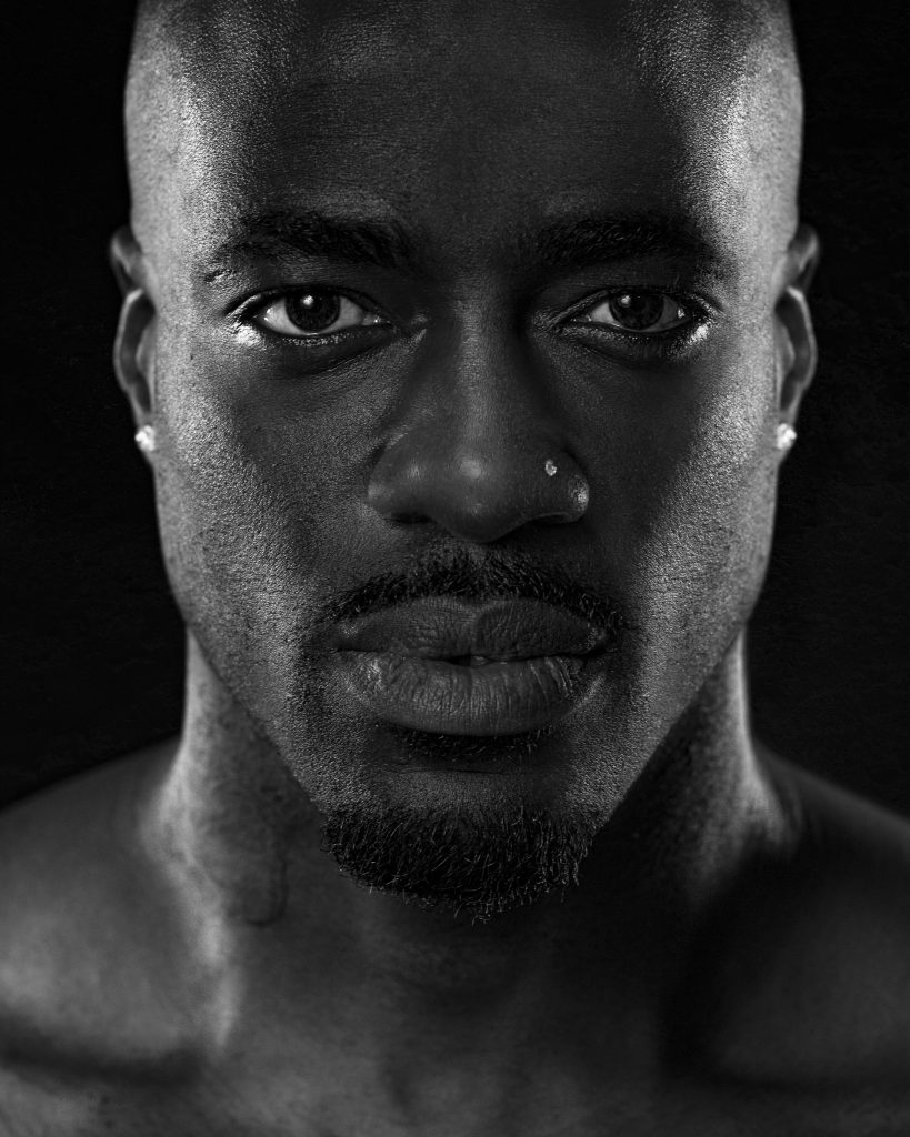 Black and white close-up of a black man with facial hair, wearing earrings, and expressing a serious demeanor. His eyes and skin catch the light dramatically.