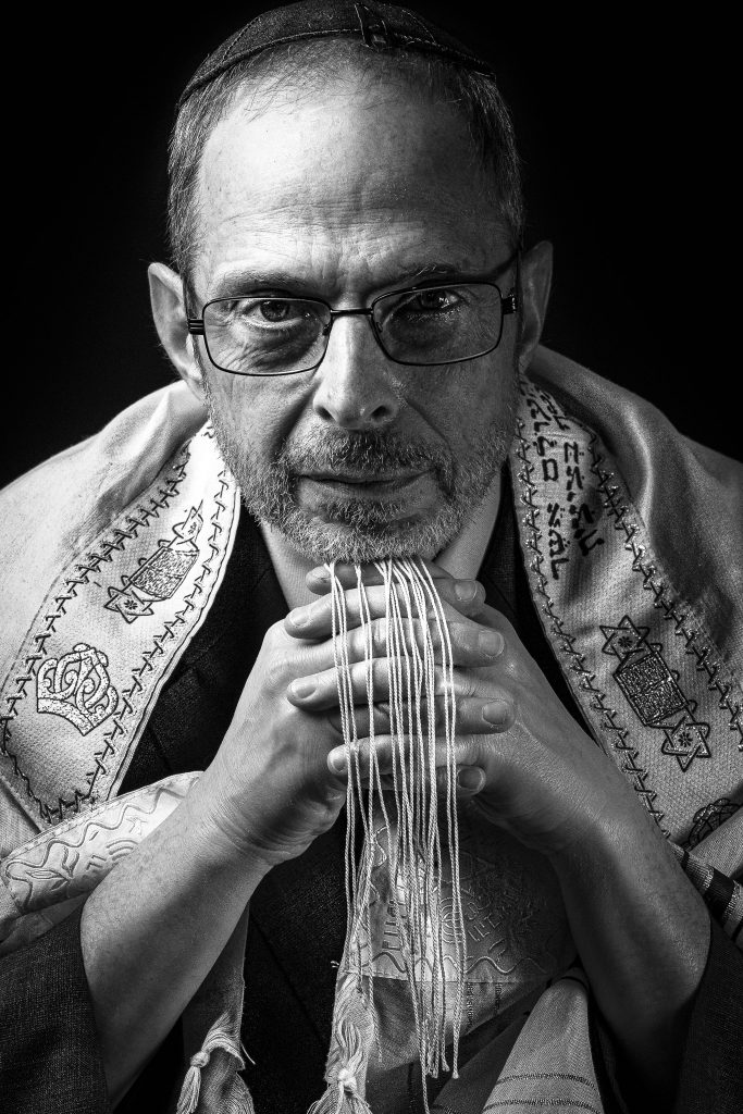 Black and white portrait of a man wearing glasses, a kippah, and a tallit, with fingers interlocked, conveying a sense of contemplation and spirituality.