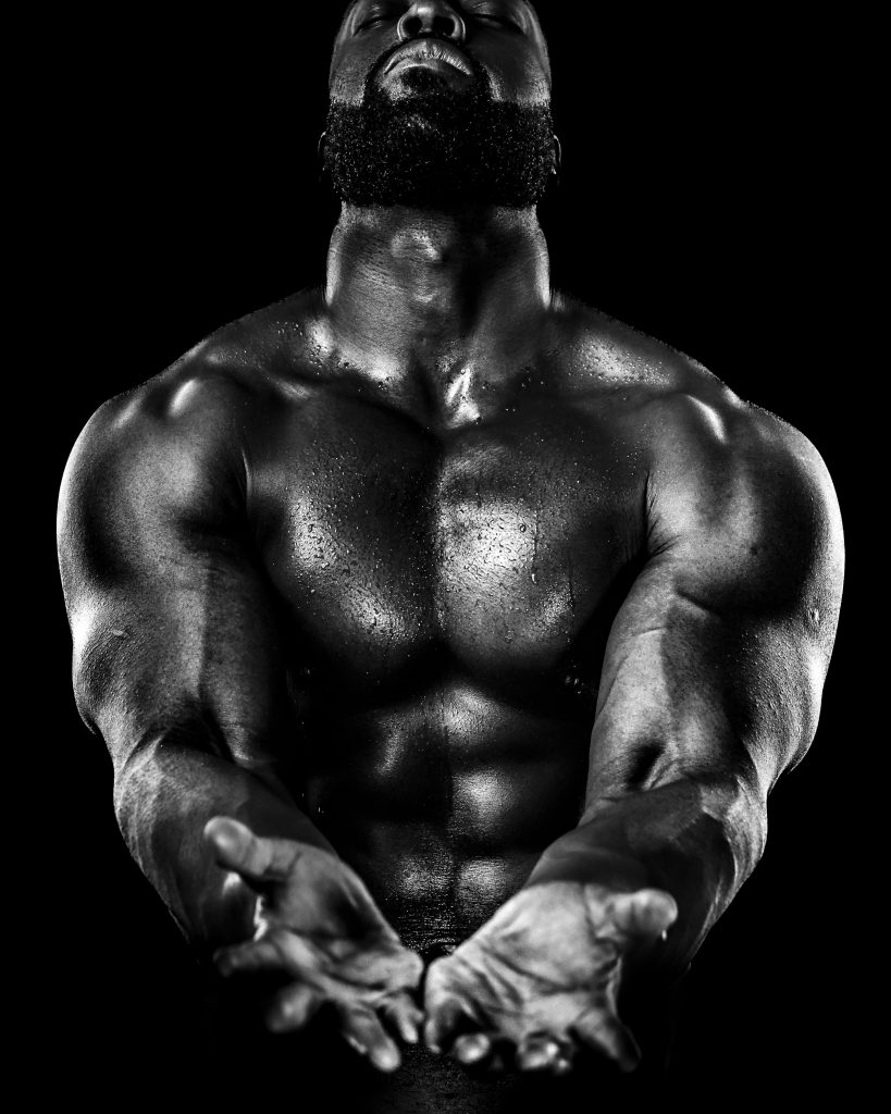 Muscular man with a beard in dramatic lighting, his bare chest glistening with sweat. He extends his hands forward, conveying strength and intensity.